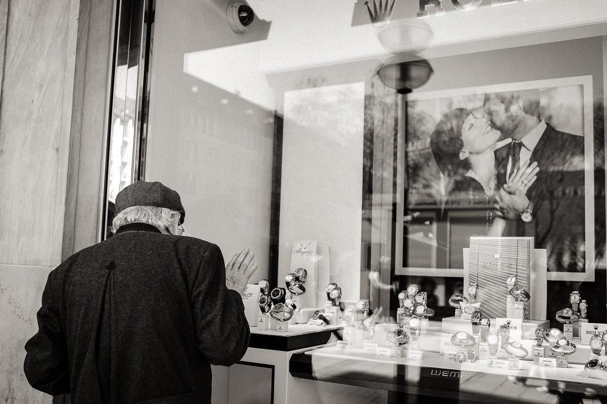 Old man looks into shop window containing watches and an image of a younger couple
